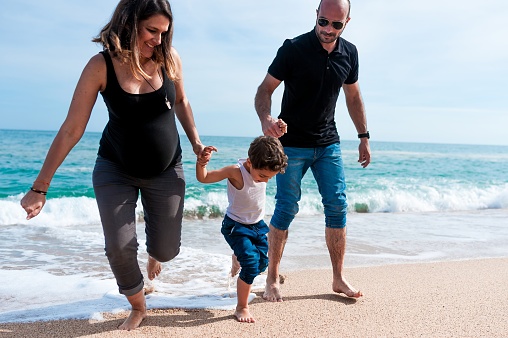 A pregnant mom and her husband holding the hands of their toddler playing at the sea.