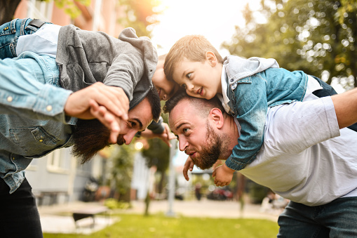 Two dads play airplane fighting with their young sons on their backs in a park. 