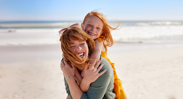 A laughing redheaded mom giving a piggback to her redheaded duaghter at the beach.