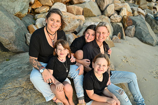 Two moms sitting on tsome rocks at the beach with their three daughters.