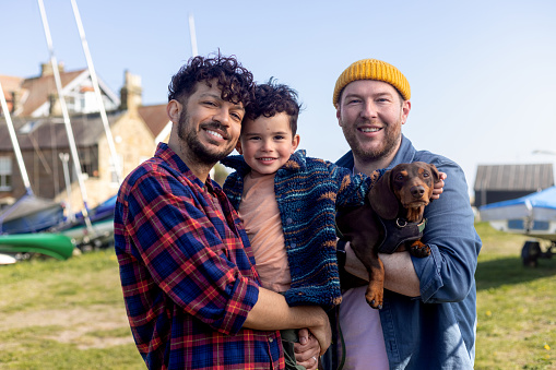 Two smiling dads holding their smiling toddler son and their dog with small boats in the background. 